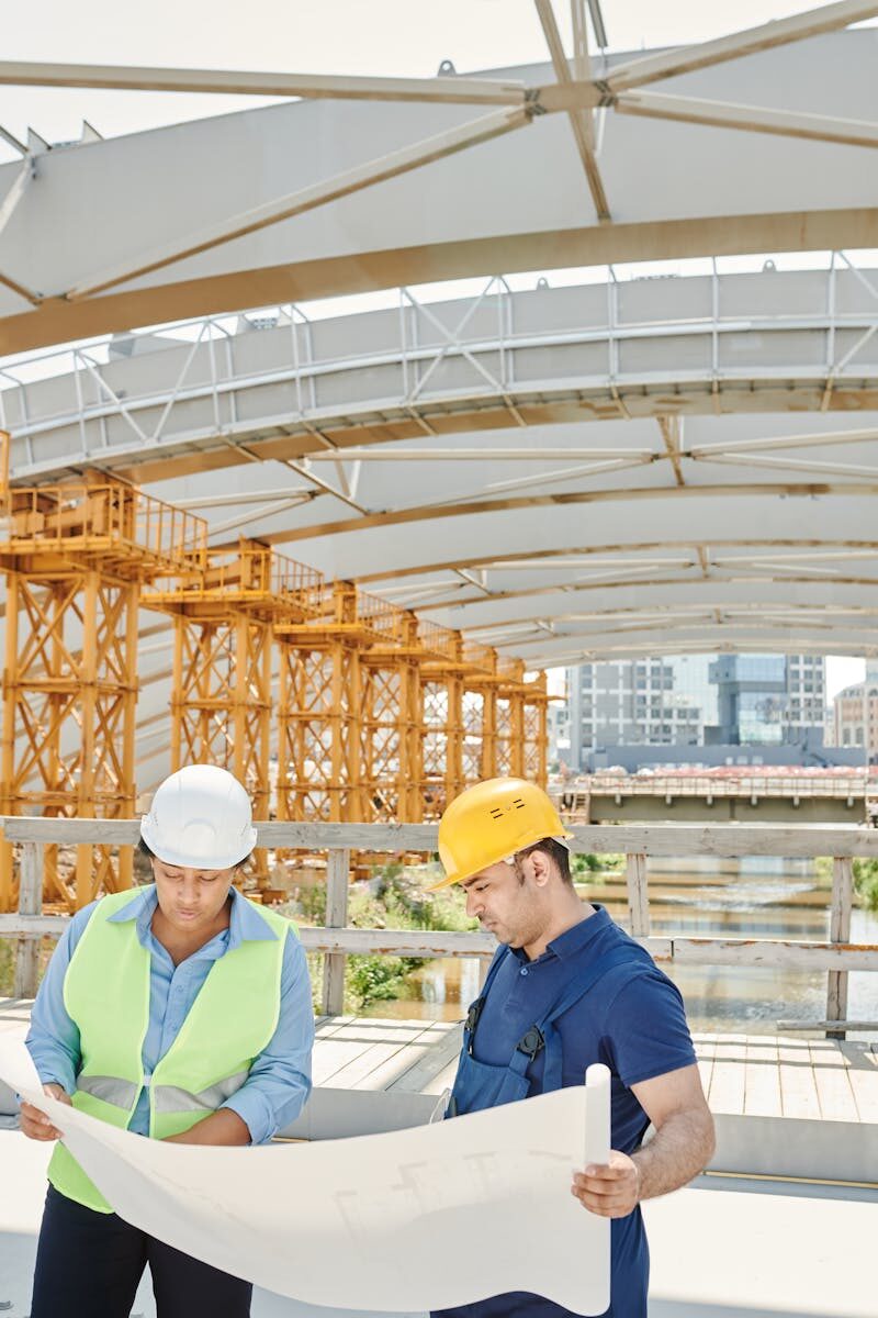 Progress Update 5 Two engineers on a construction site examining blueprints with steel structures in the background.